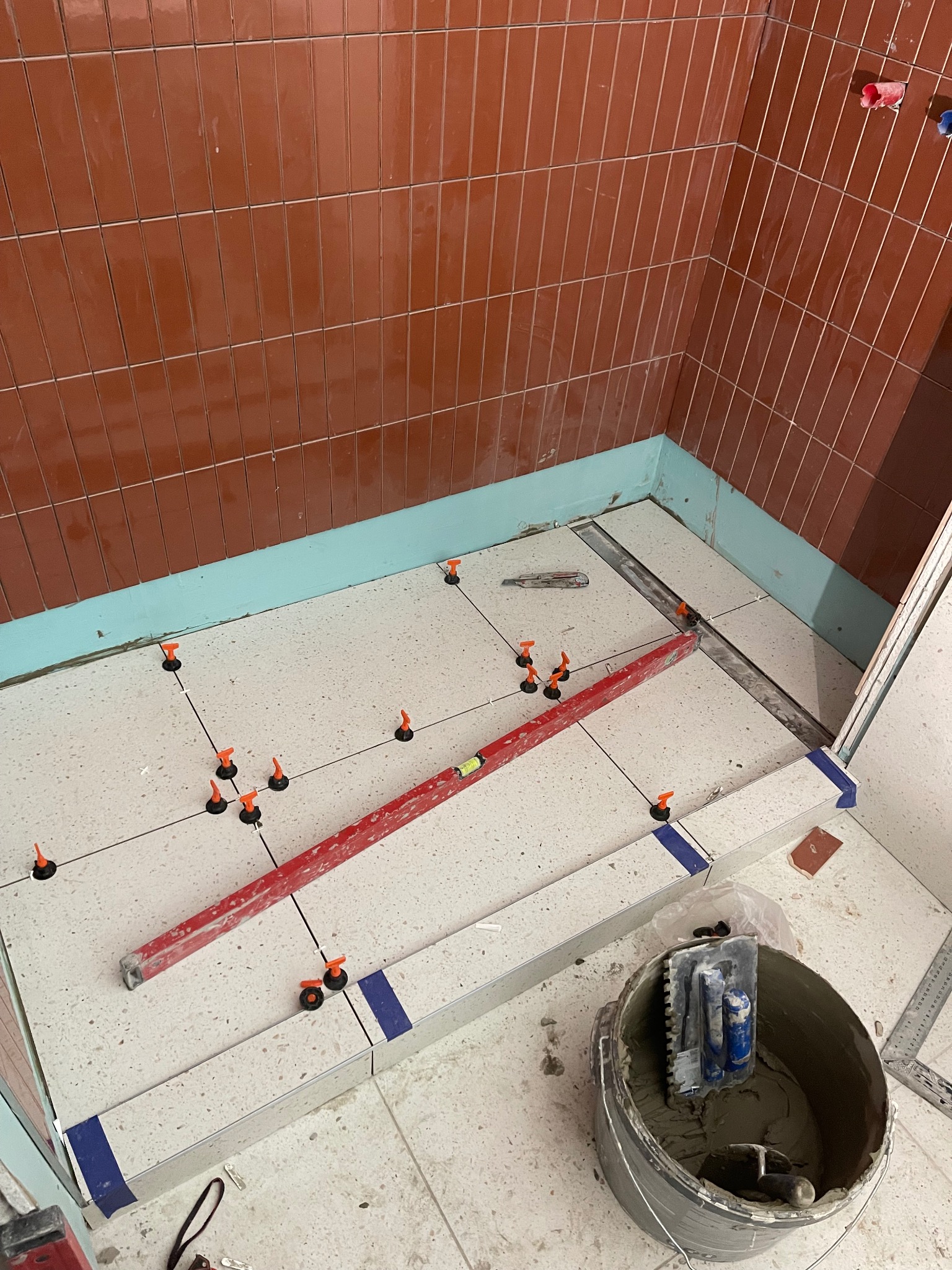 Montaż płytek podłogowych w łazience, widoczne poziomica, krzyżyki dystansowe i wiadro z zaprawą, ściany wyłożone wąskimi, brązowymi płytkami.