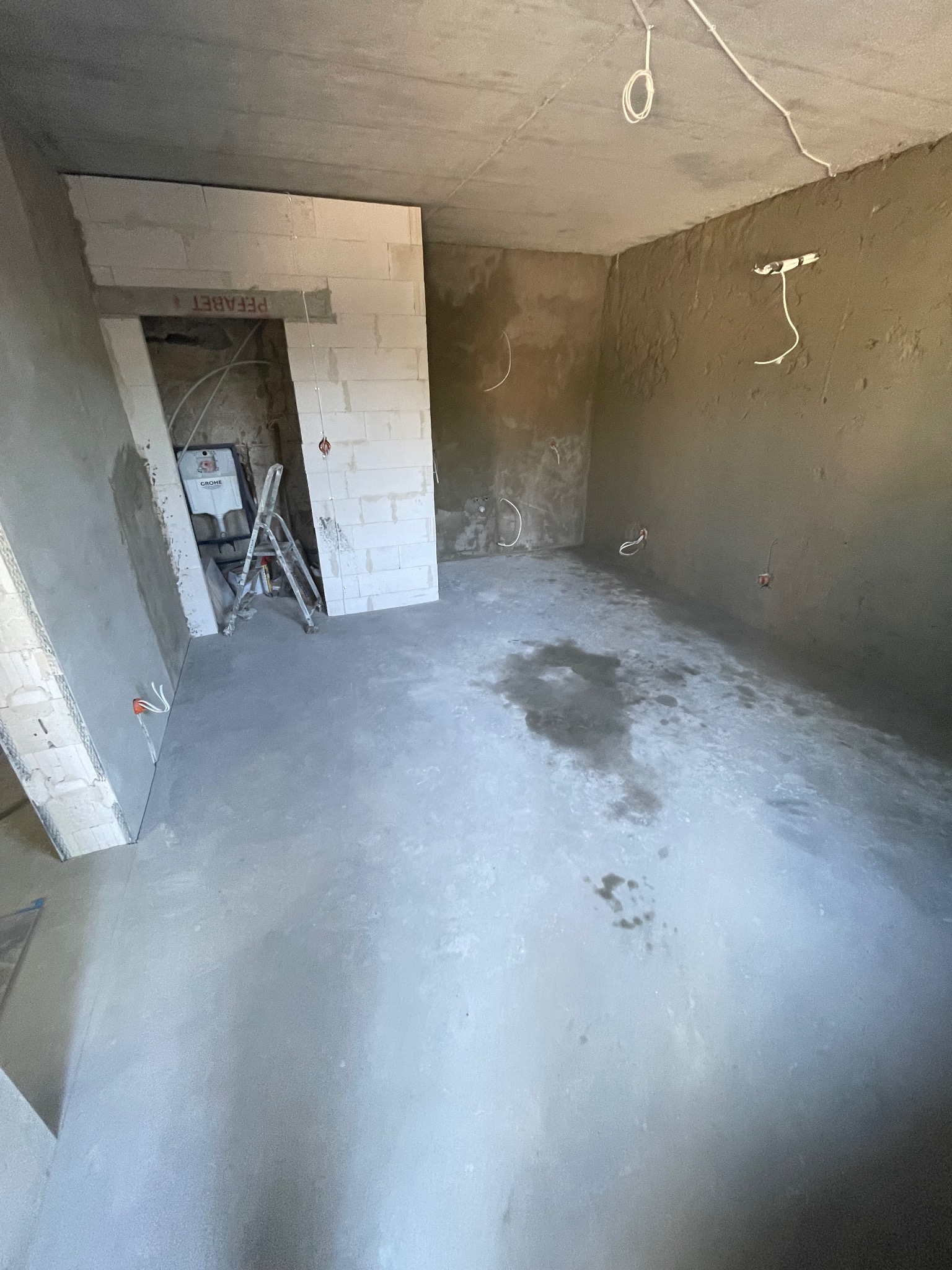 Surowy stan łazienki w Warszawie: wylewka, ściany z betonu komórkowego i instalacje elektryczne przed wykończeniem. Widoczna drabina i odpływ Grohe.