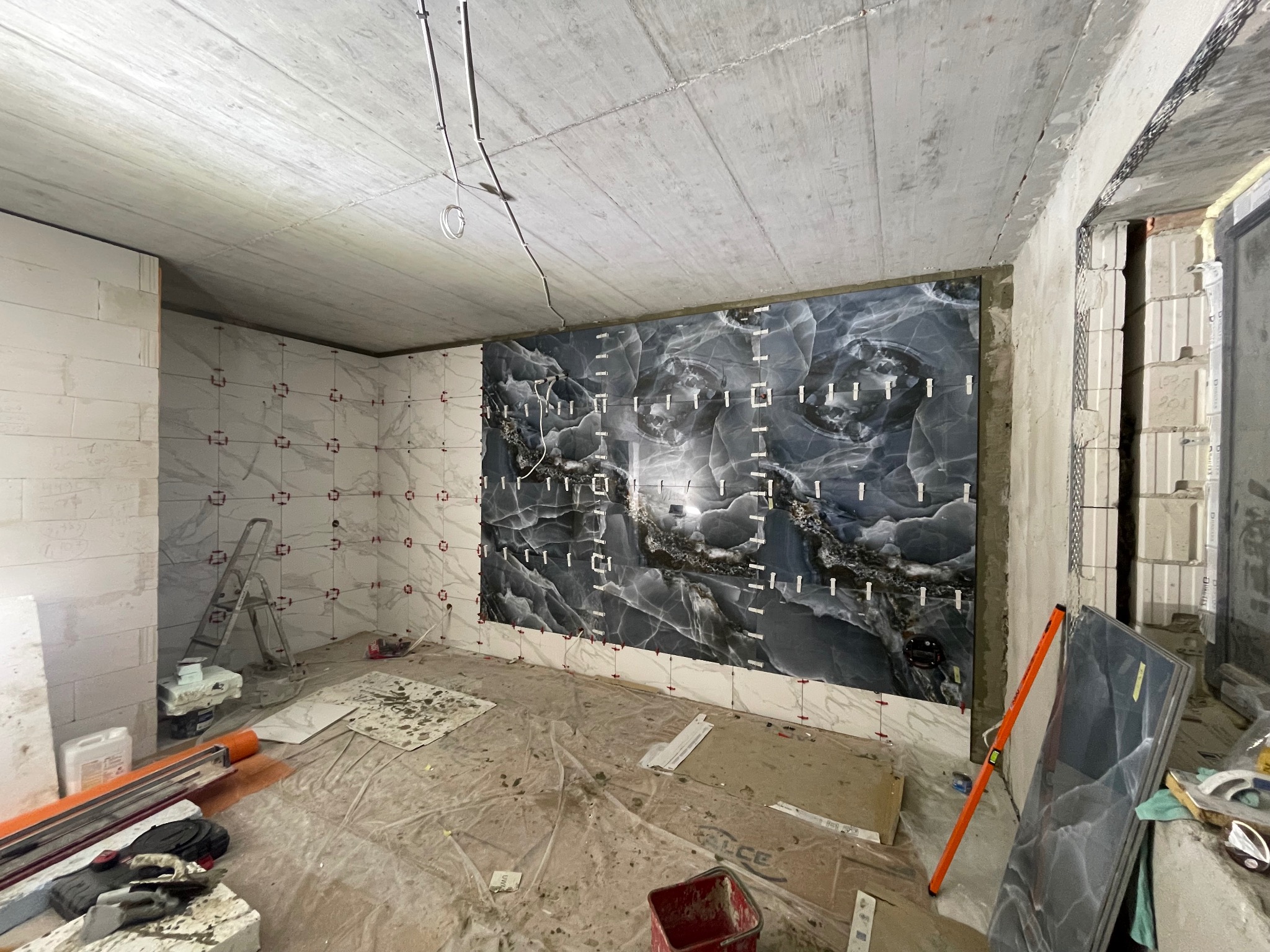 Wnętrze w trakcie remontu: ściany częściowo pokryte białymi i czarnymi płytkami imitującymi marmur, widoczne narzędzia i drabina. Surowy betonowy sufit z instalacją.
