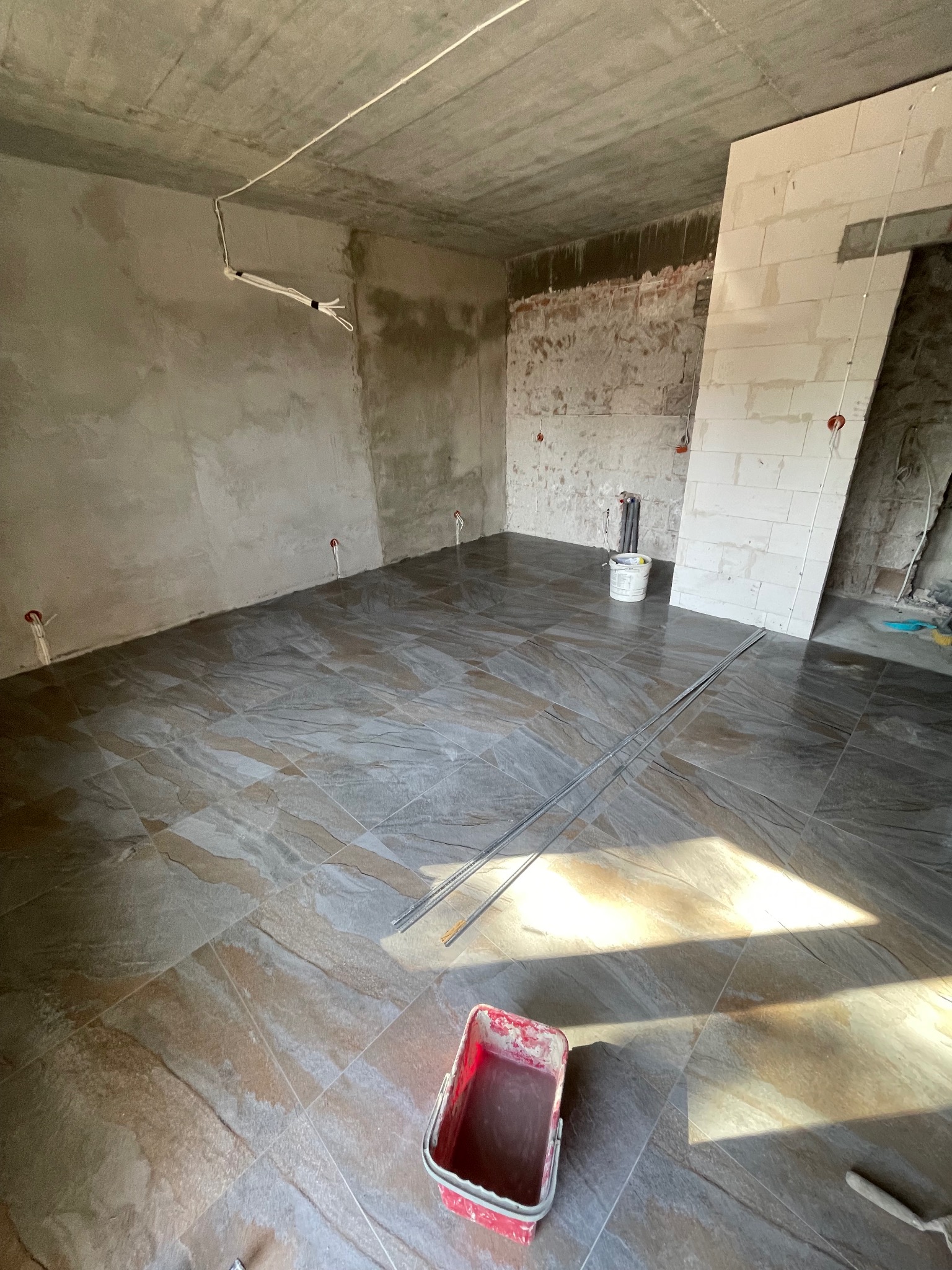 Ułożona, brązowo-szara podłoga z płytek w surowym wnętrzu. Widoczny kubeł i listwy. Ściany częściowo otynkowane, sufit betonowy.