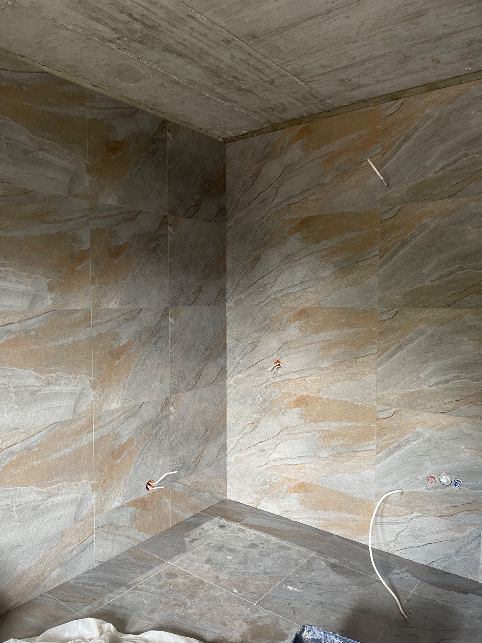 Wykończona łazienka z płytkami imitującymi kamień na ścianach i podłodze. Widoczne wystające przewody i instalacja wodna. Surowy betonowy sufit.