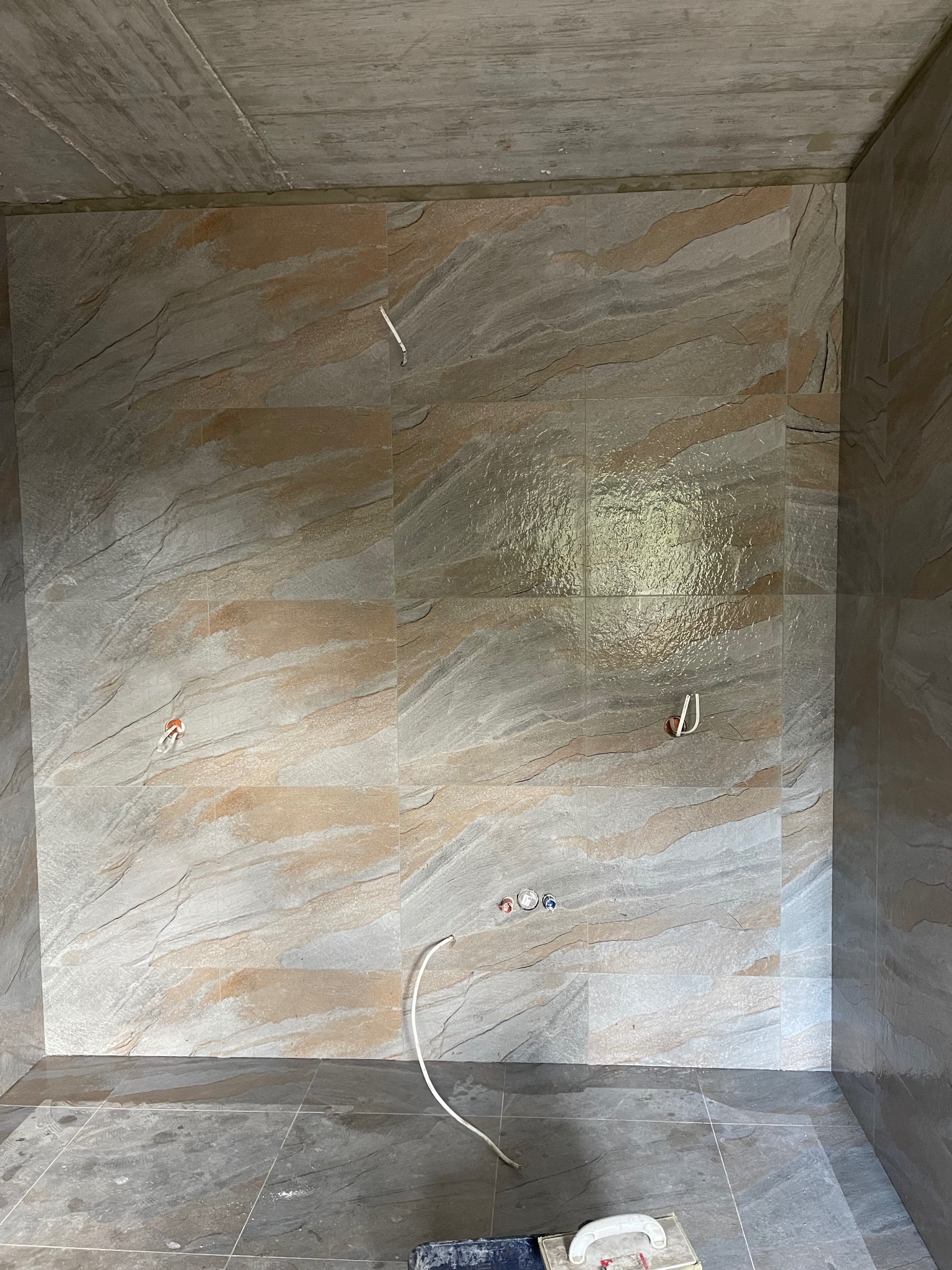 Wykończona łazienka z płytkami imitującymi kamień, widoczne wyprowadzenia instalacji wodnej i elektrycznej. Na podłodze narzędzia glazurnicze.