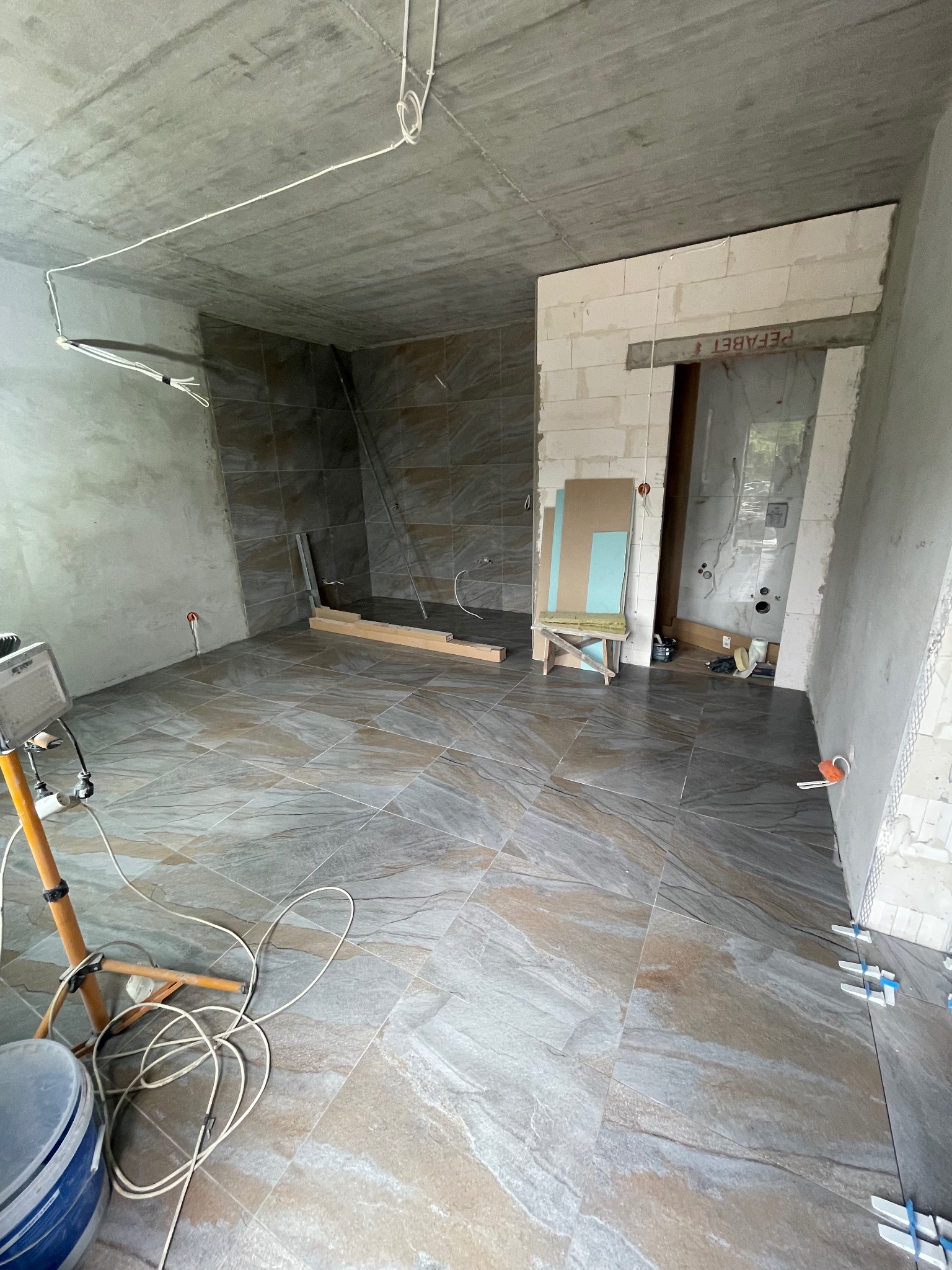 Układanie płytek podłogowych w łazience z widocznym oświetleniem roboczym, krzyżykami dystansowymi i częściowo wykończonymi ścianami.