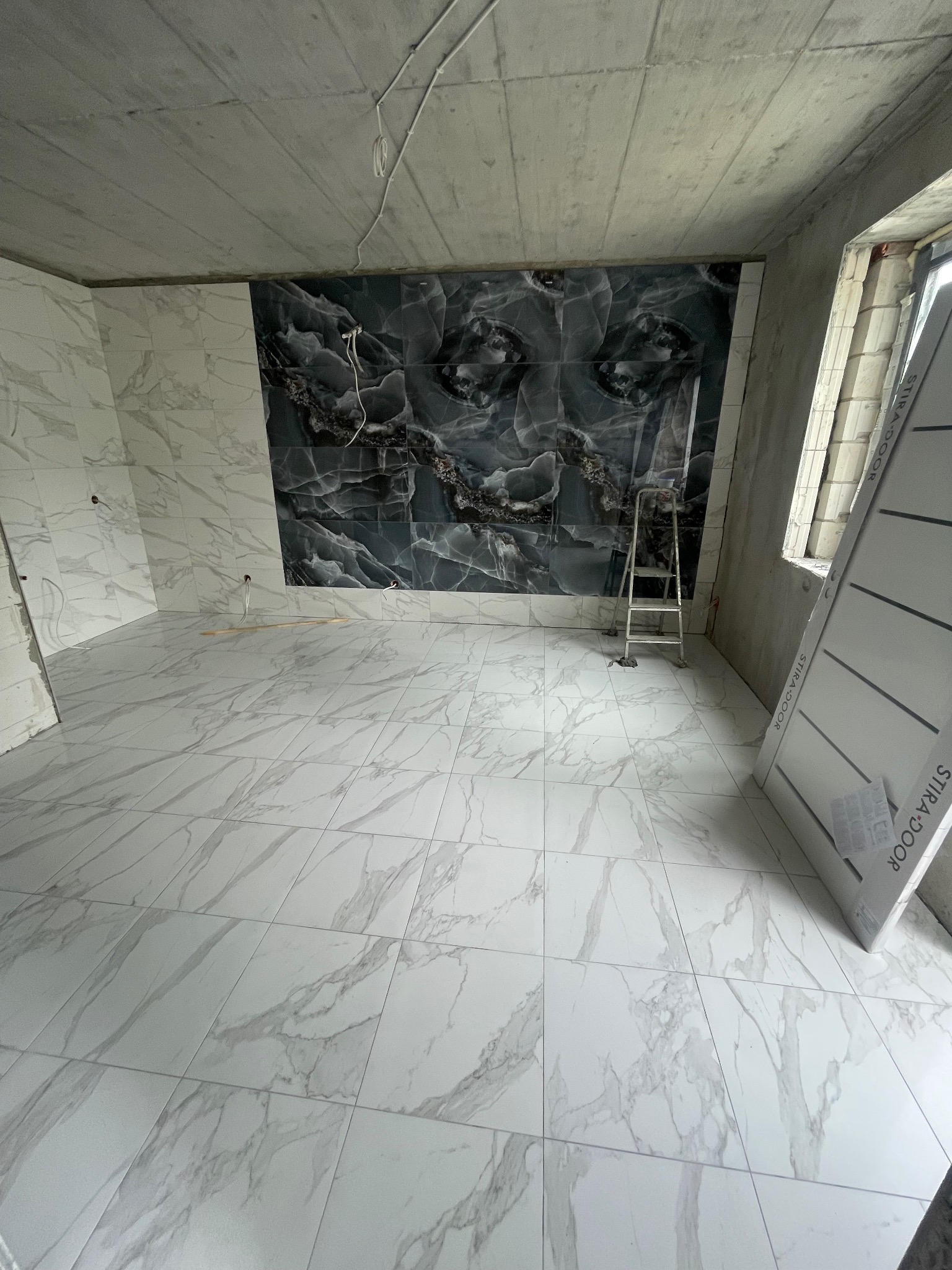 Płytki marmurowe na podłodze i ścianie z czarnym akcentem, drabina w rogu, surowy betonowy sufit, drzwi opierają się o ścianę. Wnętrze w trakcie wykańczania.