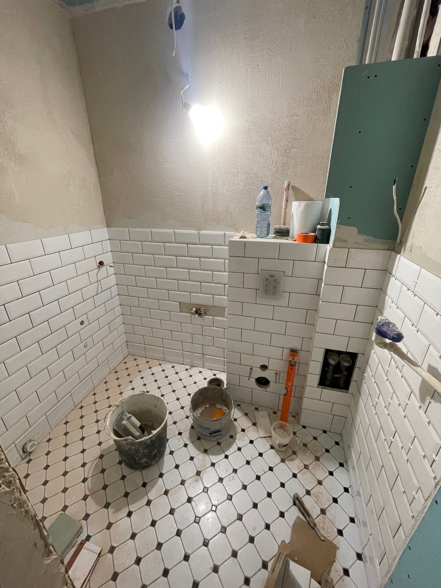 Łazienka w trakcie remontu: białe, prostokątne płytki na ścianach, wzorzysta podłoga, widoczne narzędzia i materiały budowlane, surowy stan wykończenia.