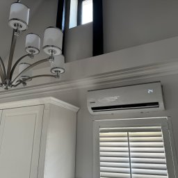 SOLFAN SPÓŁKA Z OGRANICZONĄ ODPOWIEDZIALNOŚCIĄ - Biały klimatyzator marki Kaisai zamontowany nad oknem z żaluzjami w nowoczesnym wnętrzu, obok szafki kuchennej i pod ozdobnym oświetleniem sufitowym.