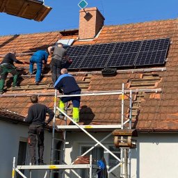 SOLFAN SPÓŁKA Z OGRANICZONĄ ODPOWIEDZIALNOŚCIĄ - Montaż paneli słonecznych na dachu z czerwonej dachówki. Czterech monterów na rusztowaniu i na dachu. Jeden z nich w niebieskim kombinezonie i jaskrawożółtych spodniach. Dach częściowo zdjęty,...