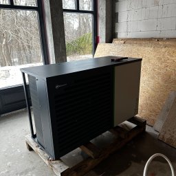 SOLFAN SPÓŁKA Z OGRANICZONĄ ODPOWIEDZIALNOŚCIĄ - Nowoczesna, czarno-biała jednostka grzewcza Heitztechnik, model Calla Verde, umieszczona na drewnianych legarach w surowym wnętrzu budynku w trakcie budowy, z widocznymi oknami i nieotynkowanymi...