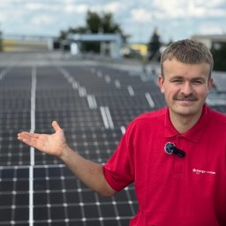 SOLFAN SPÓŁKA Z OGRANICZONĄ ODPOWIEDZIALNOŚCIĄ - Mężczyzna w czerwonej koszulce z logo firmy prezentuje instalację paneli słonecznych na dachu budynku.