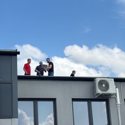 SOLFAN SPÓŁKA Z OGRANICZONĄ ODPOWIEDZIALNOŚCIĄ - Czterech mężczyzn na dachu budynku z klimatyzatorami, jeden z laptopem, błękitne niebo z chmurami odbija się w oknach.