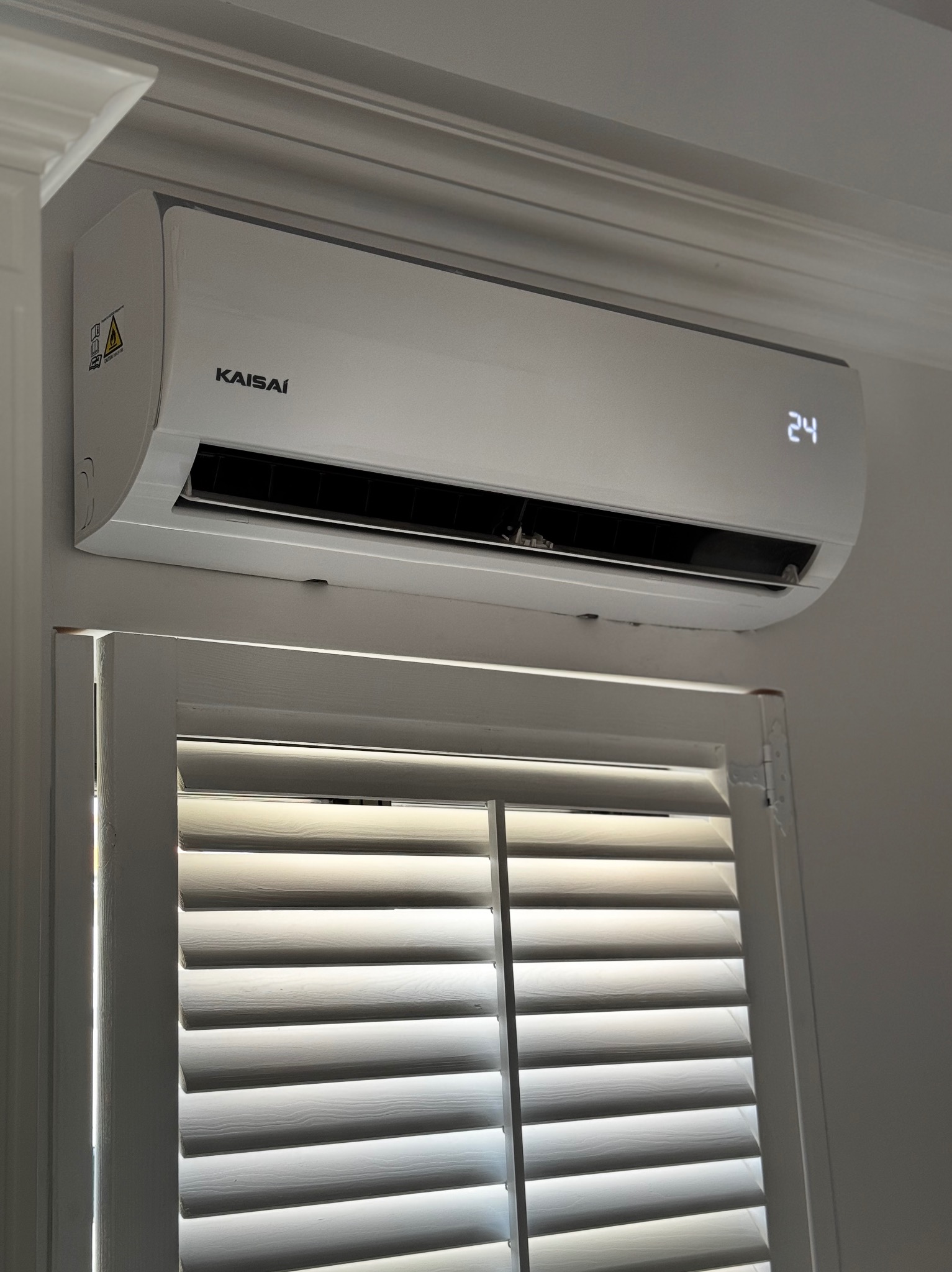 Biały klimatyzator Kaisai zamontowany nad oknem z żaluzjami w Olsztynie, wyświetlający temperaturę 24 stopnie Celsjusza.