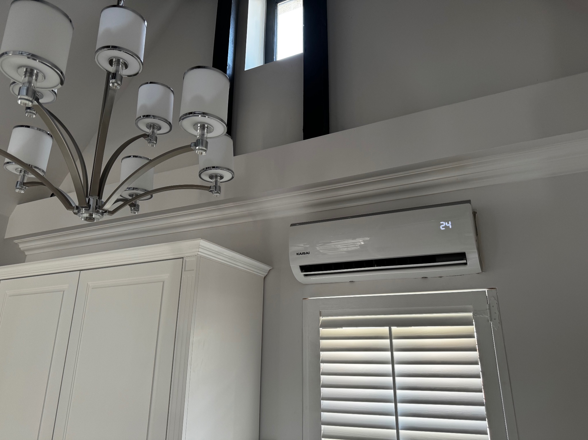 Biały klimatyzator marki Kaisai zamontowany nad oknem z żaluzjami w nowoczesnym wnętrzu, obok szafki kuchennej i pod ozdobnym oświetleniem sufitowym.