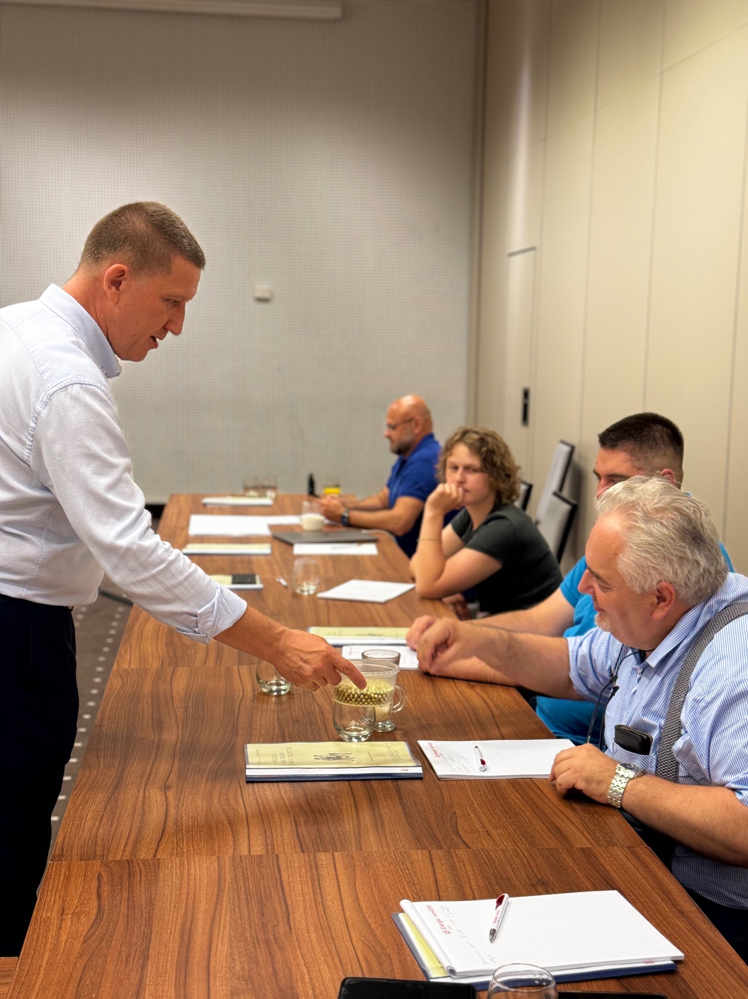 Mężczyzna w jasnej koszuli prezentuje coś na stole, wskazując palcem na szklankę z drobnymi elementami, podczas gdy inni uczestnicy siedzą przy stole i notują.