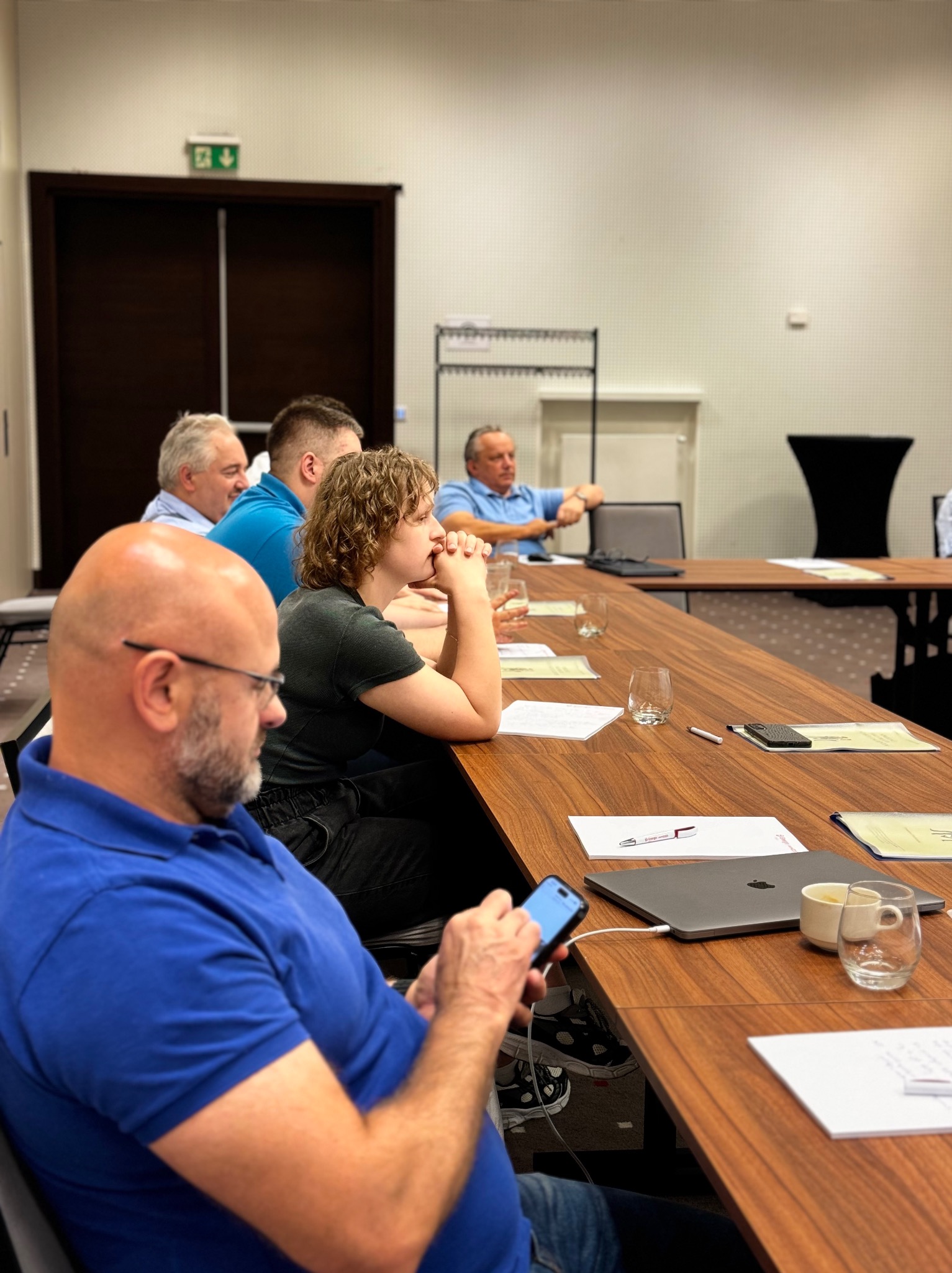 Grupa osób w sali konferencyjnej, siedząca przy długim drewnianym stole z laptopami, notatkami i szklankami, mężczyzna w niebieskiej koszuli sprawdza coś na smartfonie.