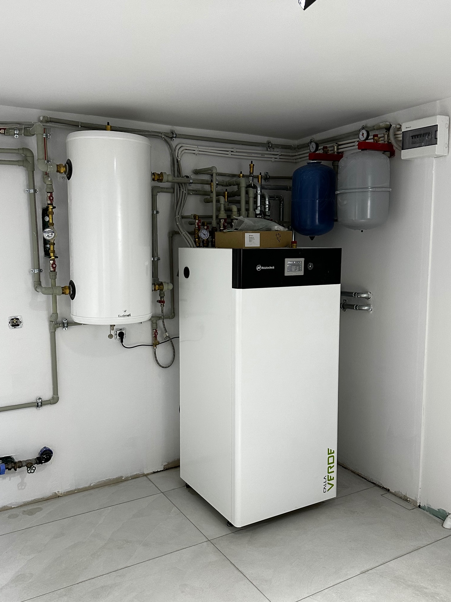 Biała kotłownia z nowoczesnym piecem grzewczym, zbiornikiem buforowym i naczyniami przeponowymi, instalacja w nowym budynku z szarymi rurami i białą skrzynką elektryczną.