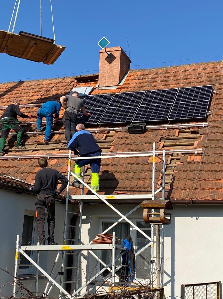 Montaż paneli słonecznych na dachu z czerwonej dachówki. Czterech monterów na rusztowaniu i na dachu. Jeden z nich w niebieskim kombinezonie i jaskrawożółtych spodniach. Dach częściowo zdjęty,...