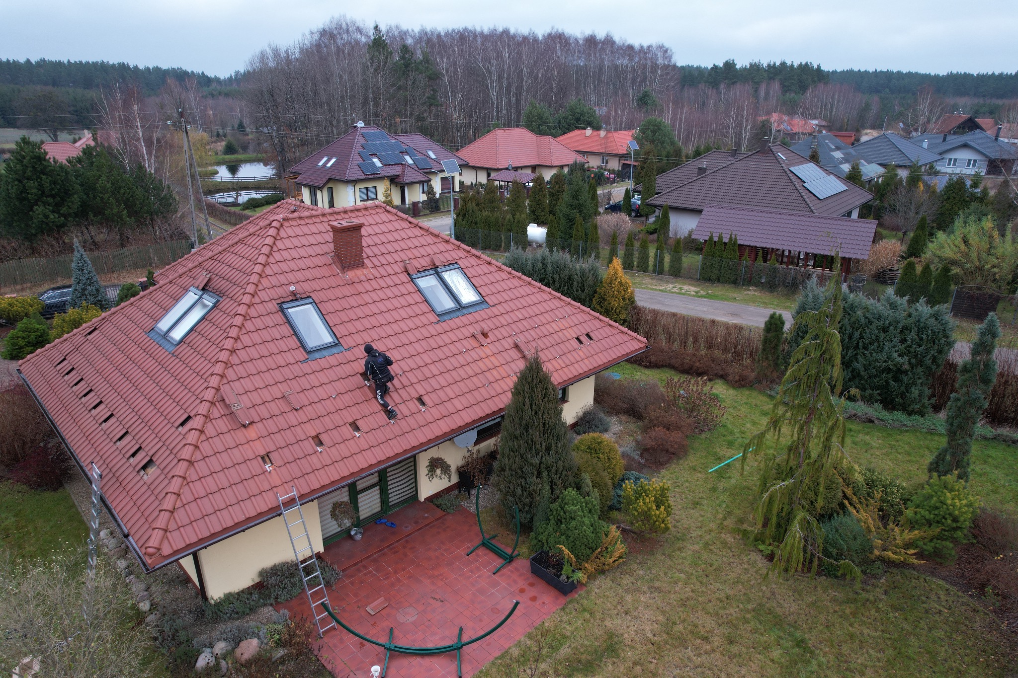 Widok z góry na dom z czerwoną dachówką, na której pracuje monter w czarnym kombinezonie, instalując panele fotowoltaiczne. W tle inne domy, niektóre z panelami słonecznymi, oraz drzewa bez liści.