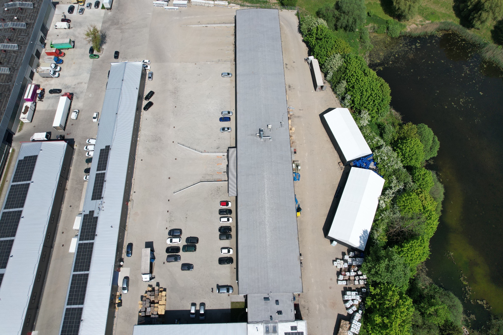 Widok z lotu ptaka na budynki przemysłowe z panelami fotowoltaicznymi na dachu, parkingiem z samochodami i dwoma białymi namiotami magazynowymi w pobliżu zieleni i zbiornika wodnego.