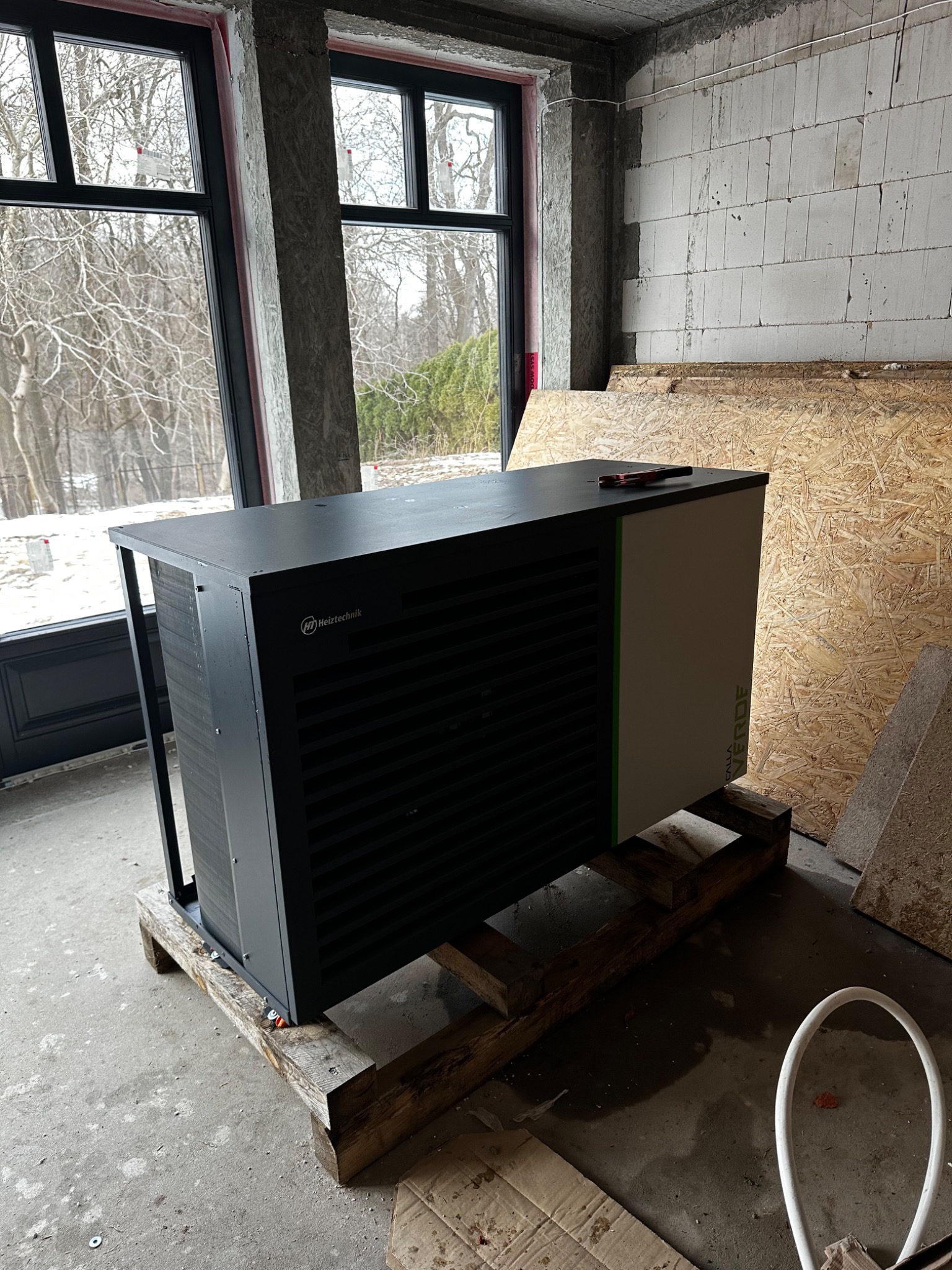 Nowoczesna, czarno-biała jednostka grzewcza Heitztechnik, model Calla Verde, umieszczona na drewnianych legarach w surowym wnętrzu budynku w trakcie budowy, z widocznymi oknami i nieotynkowanymi...