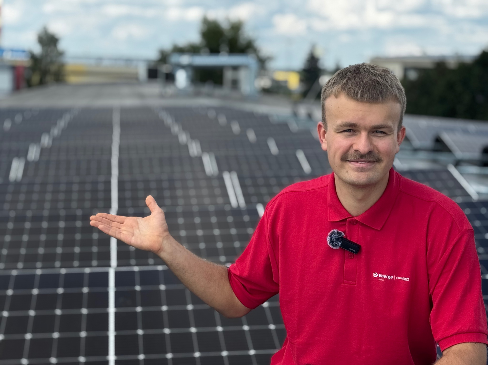 Mężczyzna w czerwonej koszulce z logo firmy prezentuje instalację paneli słonecznych na dachu budynku.