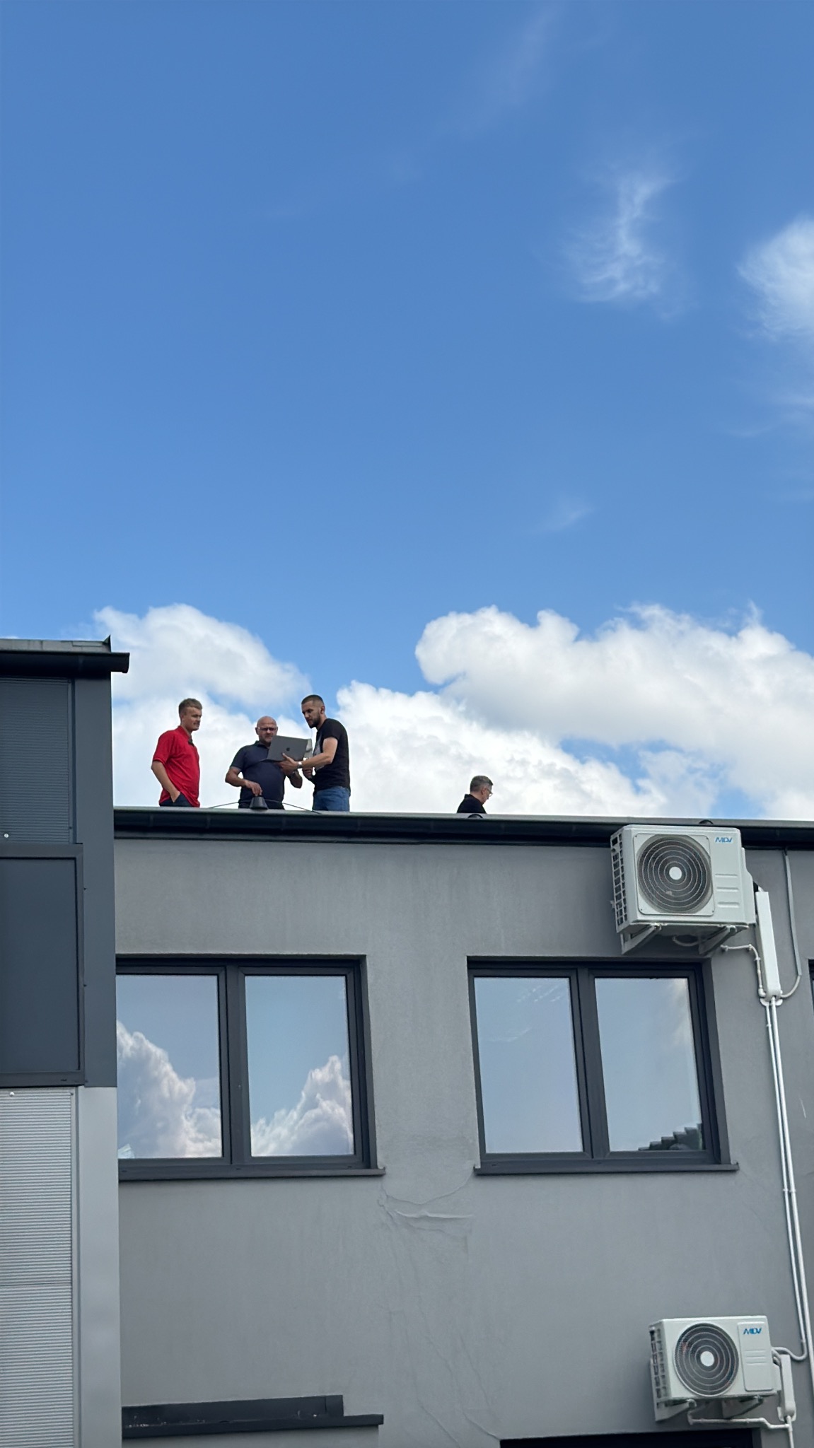 Czterech mężczyzn na dachu budynku z klimatyzatorami, jeden z laptopem, błękitne niebo z chmurami odbija się w oknach.