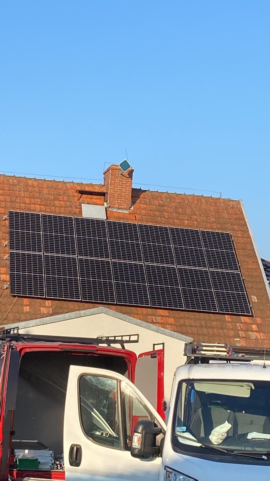 Panele słoneczne na dachu z czerwonej dachówki, widoczne dwa samochody dostawcze z otwartymi drzwiami i drabinkami dachowymi, komin z anteną na szczycie.