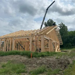 Konstrukcja szkieletowa domu w trakcie budowy, z widocznym żurawiem i pracownikiem na dachu. W tle zieleń, a w oddali przenośna toaleta.