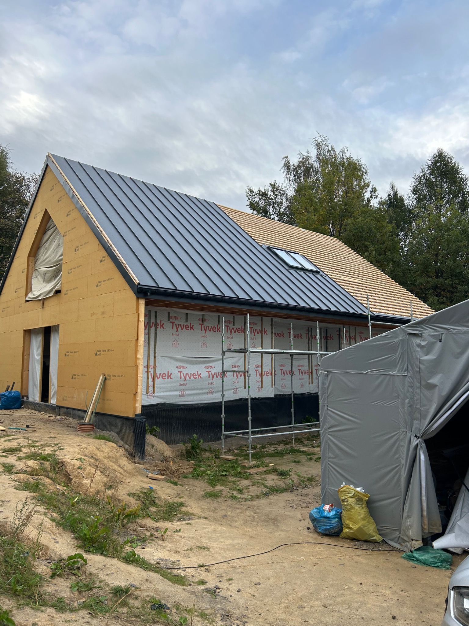 Budowa domu w stanie surowym otwartym, widoczna konstrukcja ścian z płyt OSB, częściowo pokryty blachą dach, fragment dachu pokryty drewnianą łatą, rusztowania przy ścianie szczytowej, folia...