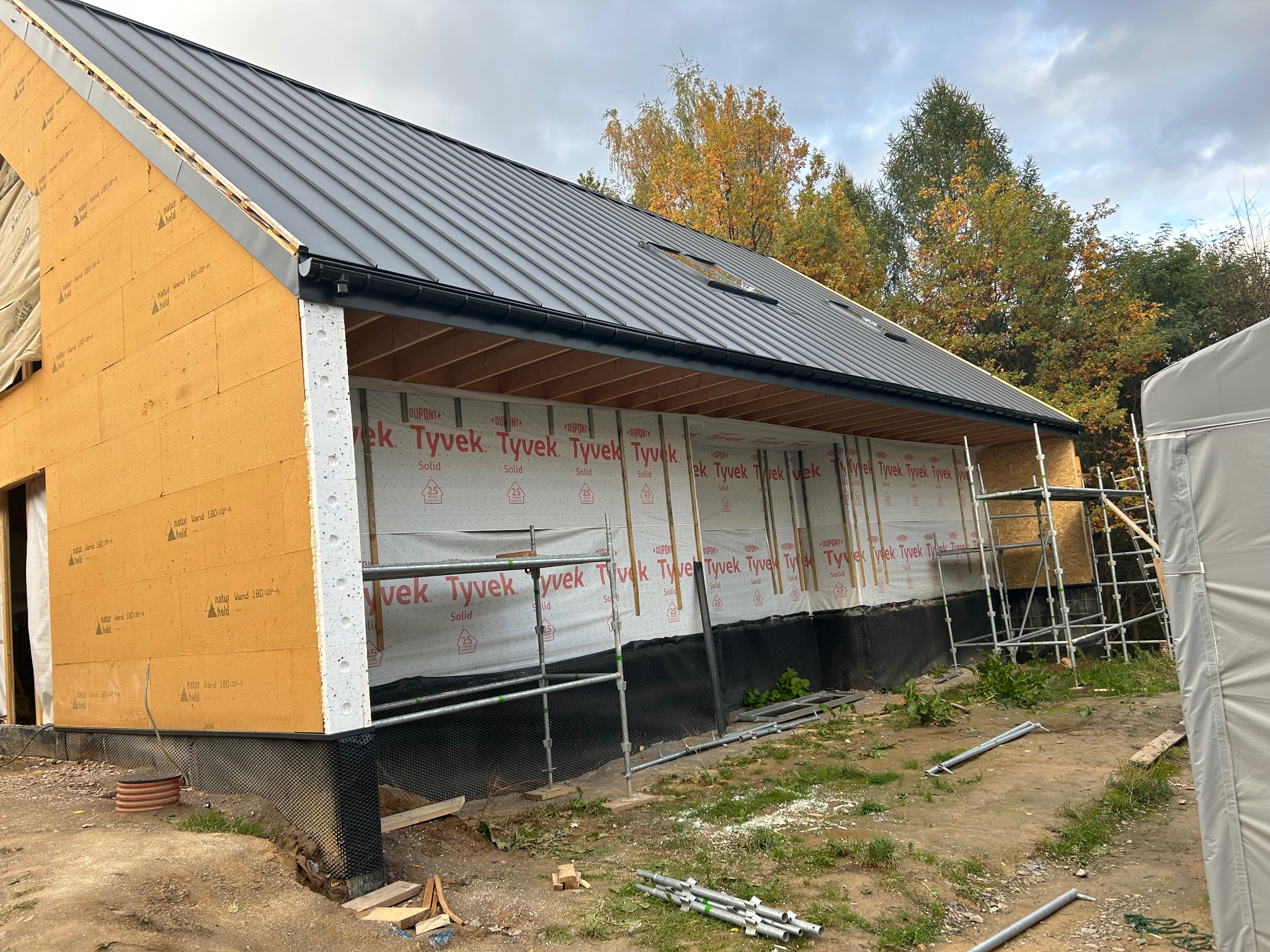 Budowa domu w stanie surowym otwartym: widoczne ściany z izolacją, czarny dach z blachy, rynna, rusztowania i folia wiatroizolacyjna Tyvek.