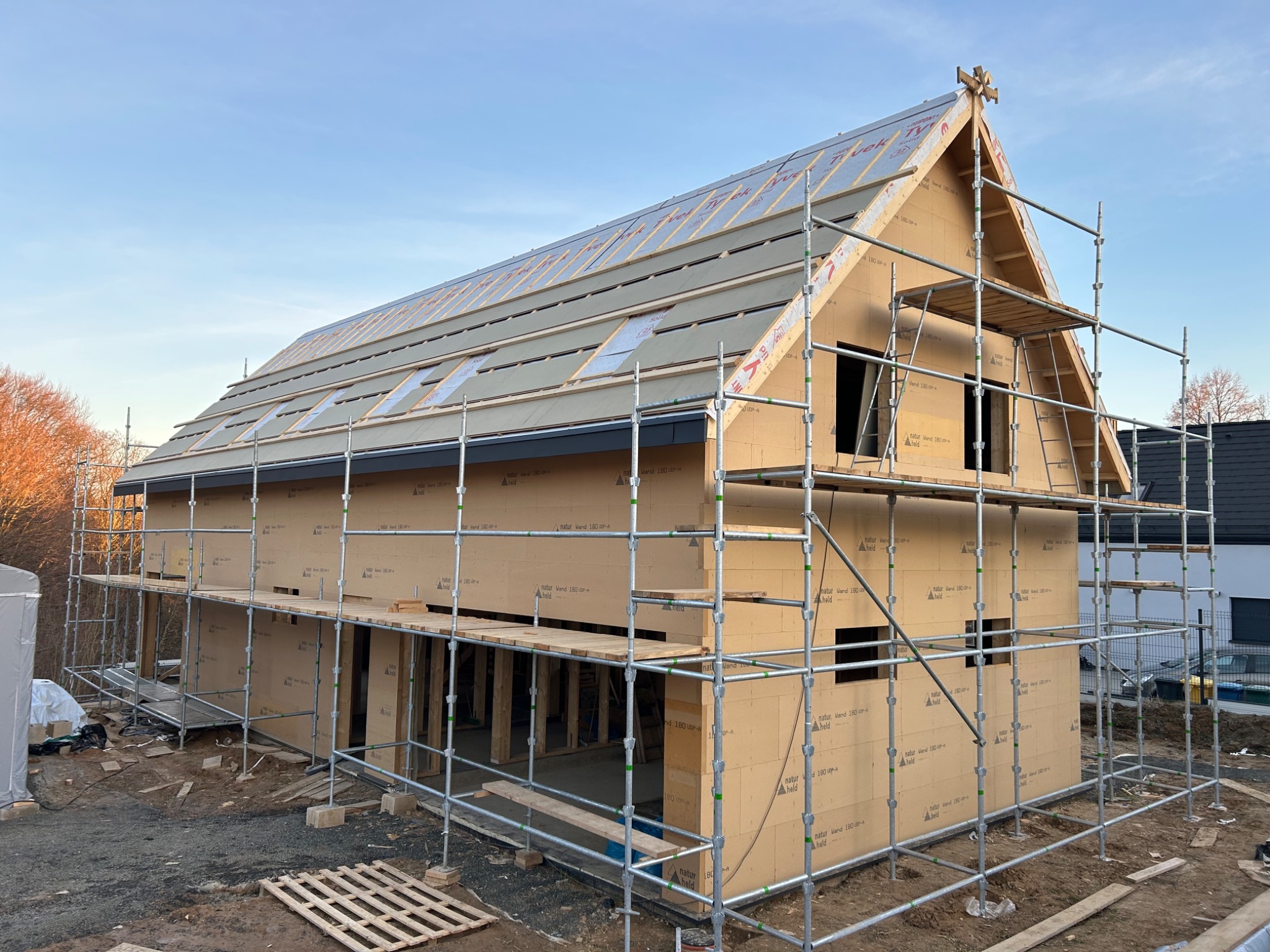 Surowy stan dwukondygnacyjnego budynku w trakcie budowy z widocznymi rusztowaniami i panelami ściennymi z jasnego drewna, z elementami dachu w trakcie montażu i ozdobnym detalem na szczycie.