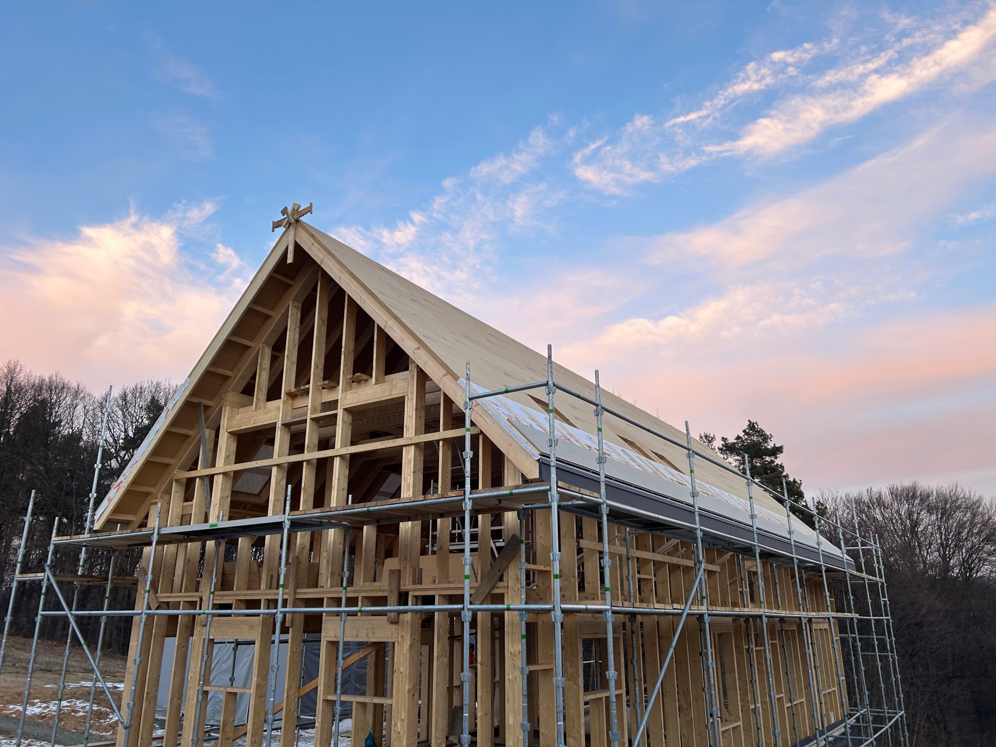 Wznoszona konstrukcja drewniana domu w technologii szkieletowej na tle nieba z pastelowymi chmurami, widoczne rusztowania i elementy wykończeniowe dachu.