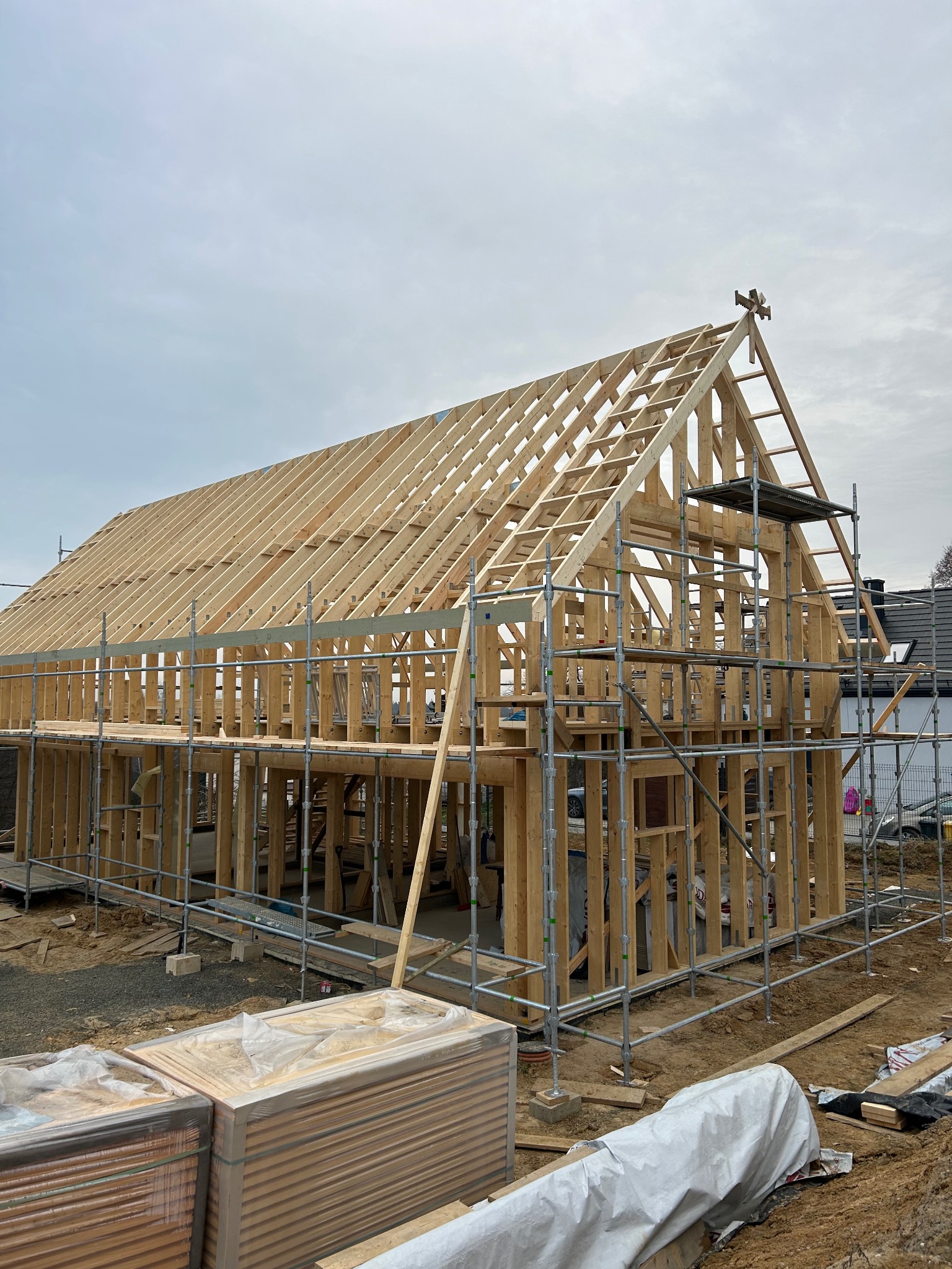 Wznoszenie konstrukcji drewnianej domu, widoczne rusztowania i elementy więźby dachowej na tle pochmurnego nieba.
