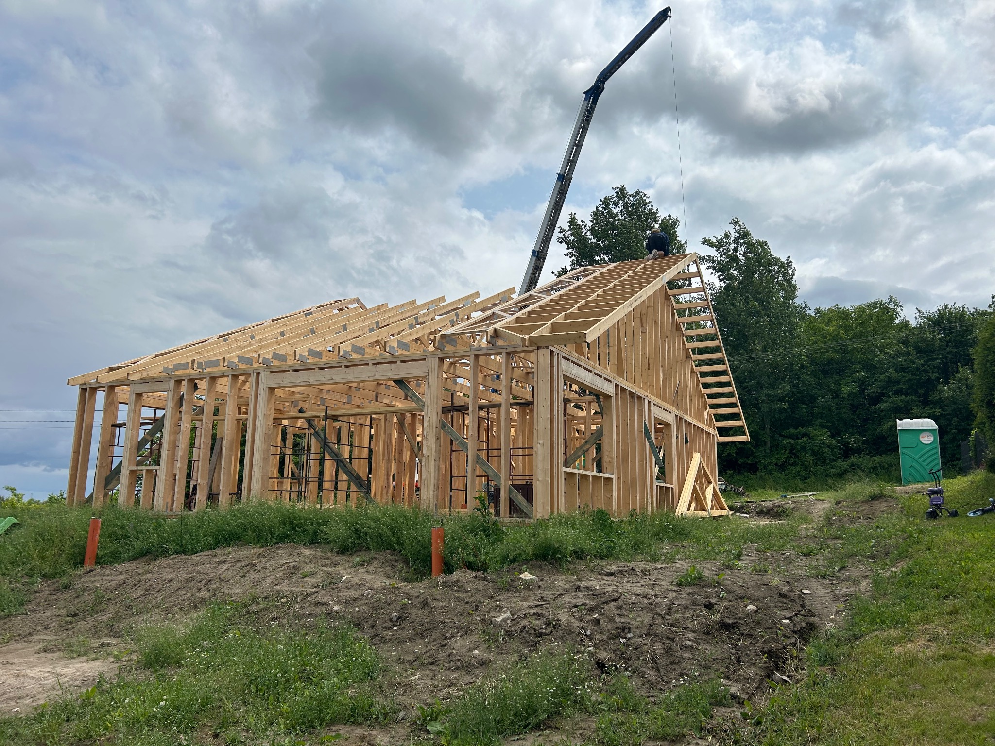 Konstrukcja szkieletowa domu w trakcie budowy, z widocznym żurawiem i pracownikiem na dachu. W tle zieleń, a w oddali przenośna toaleta.