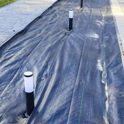 KW SPARK Usługi Elektryczne Karol Wójtowicz - Instalacja oświetlenia zewnętrznego w ogrodzie: czarne, niskie lampy z białymi kloszami zamontowane w agrowłókninie na tle domu z brązowymi oknami i drzwiami.