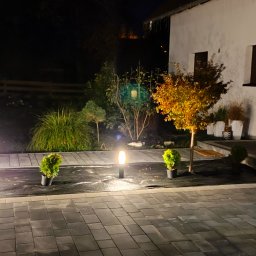 KW SPARK Usługi Elektryczne Karol Wójtowicz - Nocne oświetlenie posesji z kostki brukowej, lampy ogrodowe wzdłuż trawnika, małe drzewka w doniczkach, budynek z białymi ścianami i niebieski pojemnik na śmieci.