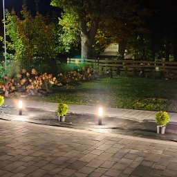 KW SPARK Usługi Elektryczne Karol Wójtowicz - Nocny widok na ogród z oświetleniem ścieżki, małe tuje w donicach i czarna folia rozłożona na trawniku, w tle dom i drewniane ogrodzenie.