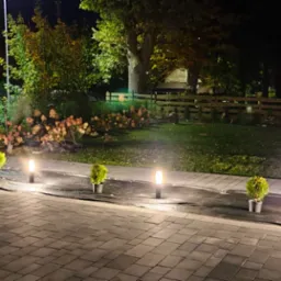 Nocny widok na ogr&oacute;d z oświetleniem ścieżki, małe tuje w donicach i czarna folia rozłożona na trawniku, w tle dom i drewniane ogrodzenie.