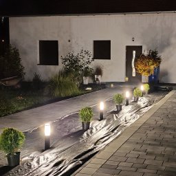 KW SPARK Usługi Elektryczne Karol Wójtowicz - Oświetlona ścieżka z kostki brukowej prowadząca do domu, z lampkami i młodymi drzewkami w donicach, zabezpieczona czarną folią przed nasadzeniami.