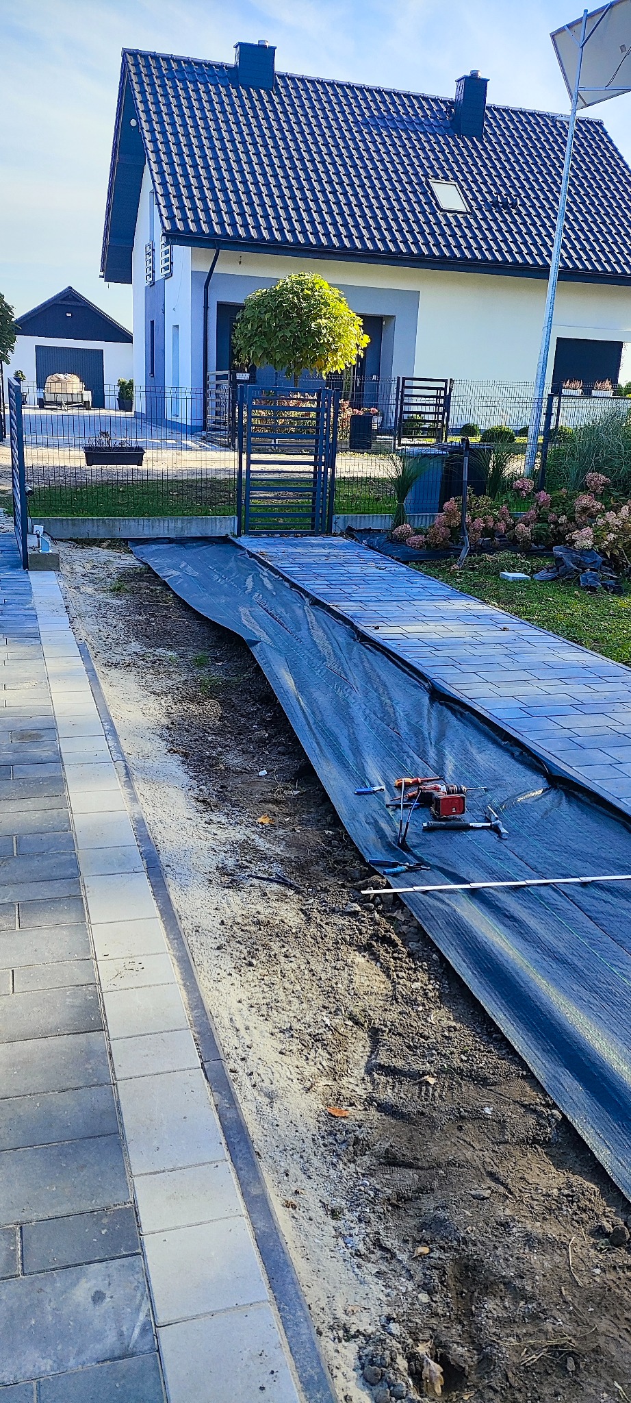 Układanie kostki brukowej na posesji przed domem, widoczne narzędzia na czarnej folii, częściowo ułożona nawierzchnia, w tle brama i dom z ciemnym dachem.