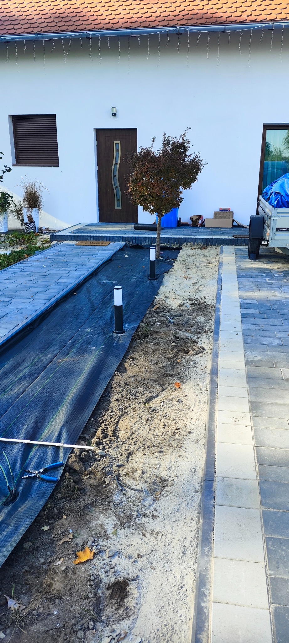 Przygotowanie terenu pod oświetlenie ogrodu: czarna agrowłóknina rozłożona wzdłuż ścieżki z kostki brukowej, widoczne lampy ogrodowe, narzędzia i przewody elektryczne.