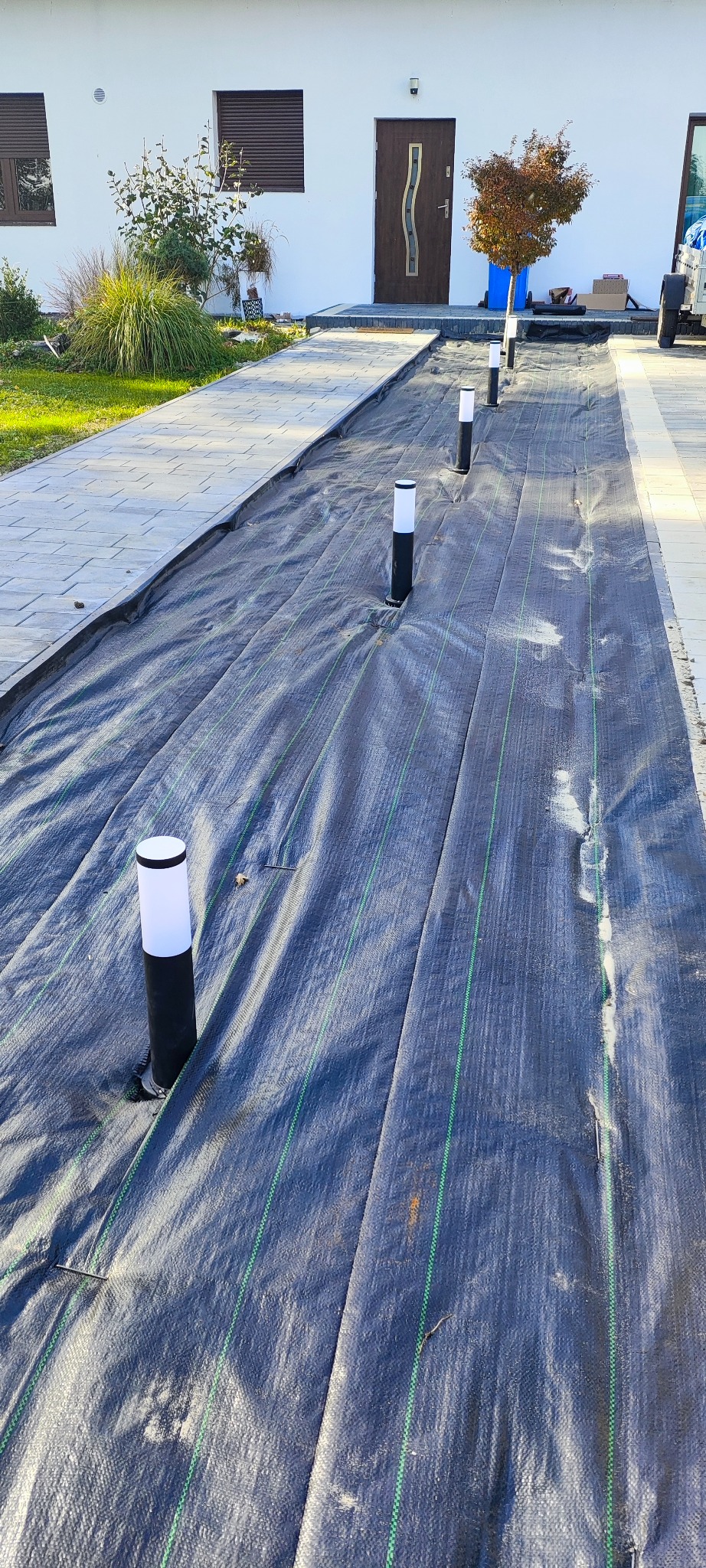 Instalacja oświetlenia zewnętrznego w ogrodzie: czarne, niskie lampy z białymi kloszami zamontowane w agrowłókninie na tle domu z brązowymi oknami i drzwiami.