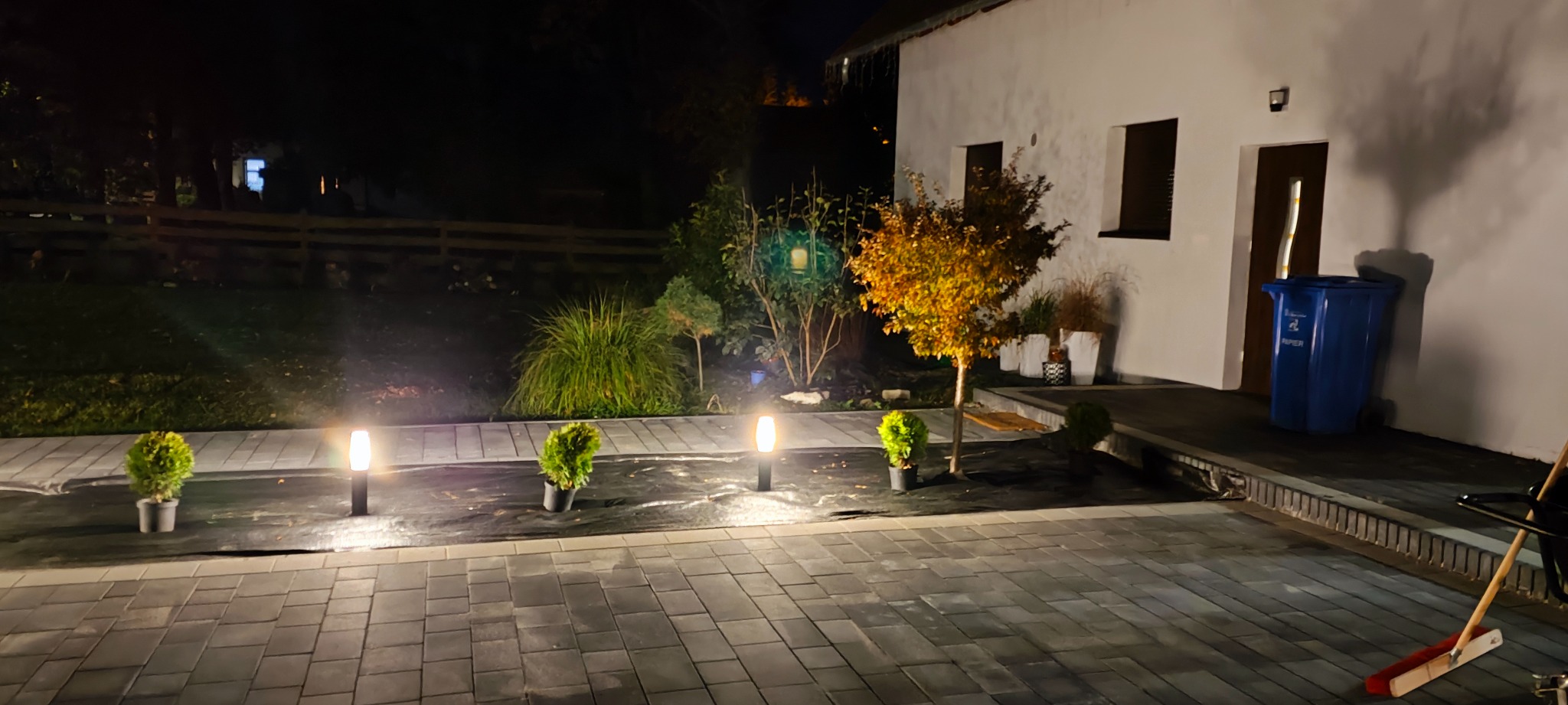 Nocne oświetlenie posesji z kostki brukowej, lampy ogrodowe wzdłuż trawnika, małe drzewka w doniczkach, budynek z białymi ścianami i niebieski pojemnik na śmieci.