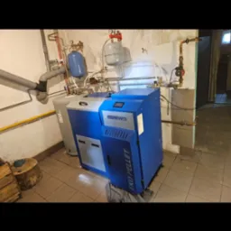 Niebiesko-szary piec EKO PELLET marki Ogniwo z podłączonymi rurami i zbiornikami w kotłowni z beżowymi płytkami. Widoczny fragment pnia drzewa.