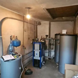 Kotłownia z piecem na paliwo stałe marki Ogniwo, zbiornikami buforowymi i osprzętem. Widoczne rury, zawory i instalacja. Na zbiorniku notatki i butelka wody.