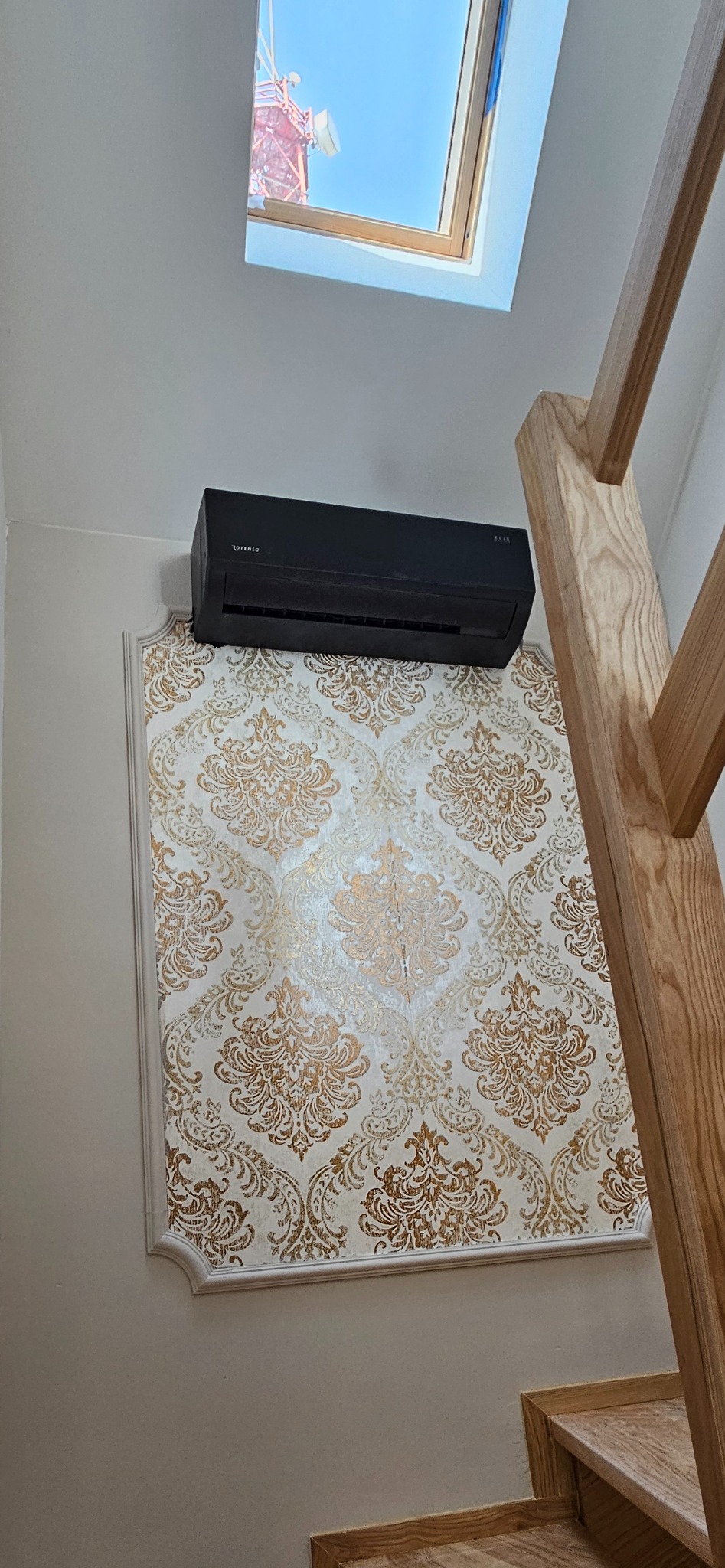 Czarny klimatyzator Rotenso na ścianie z dekoracyjną tapetą w złote wzory, widok z dołu, fragment drewnianych schodów i okno dachowe w tle.