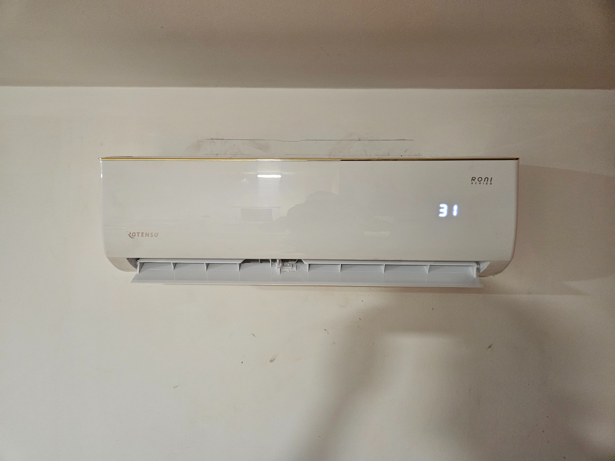 Biały klimatyzator Rotenso z wyświetlaczem temperatury (31 stopni Celsjusza) zamontowany na jasnej ścianie. Widoczne detale obudowy i żaluzje wylotu powietrza.