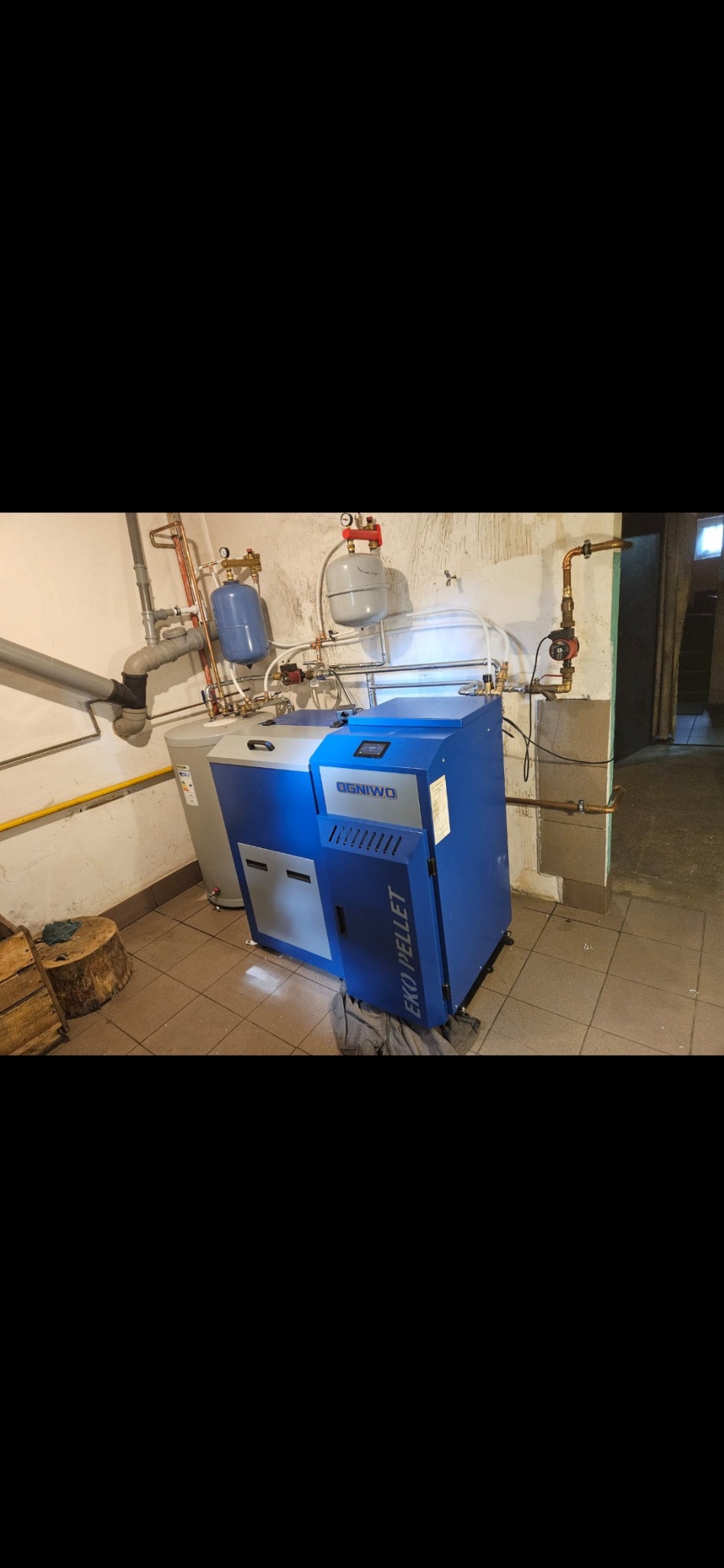 Niebiesko-szary piec EKO PELLET marki Ogniwo z podłączonymi rurami i zbiornikami w kotłowni z beżowymi płytkami. Widoczny fragment pnia drzewa.