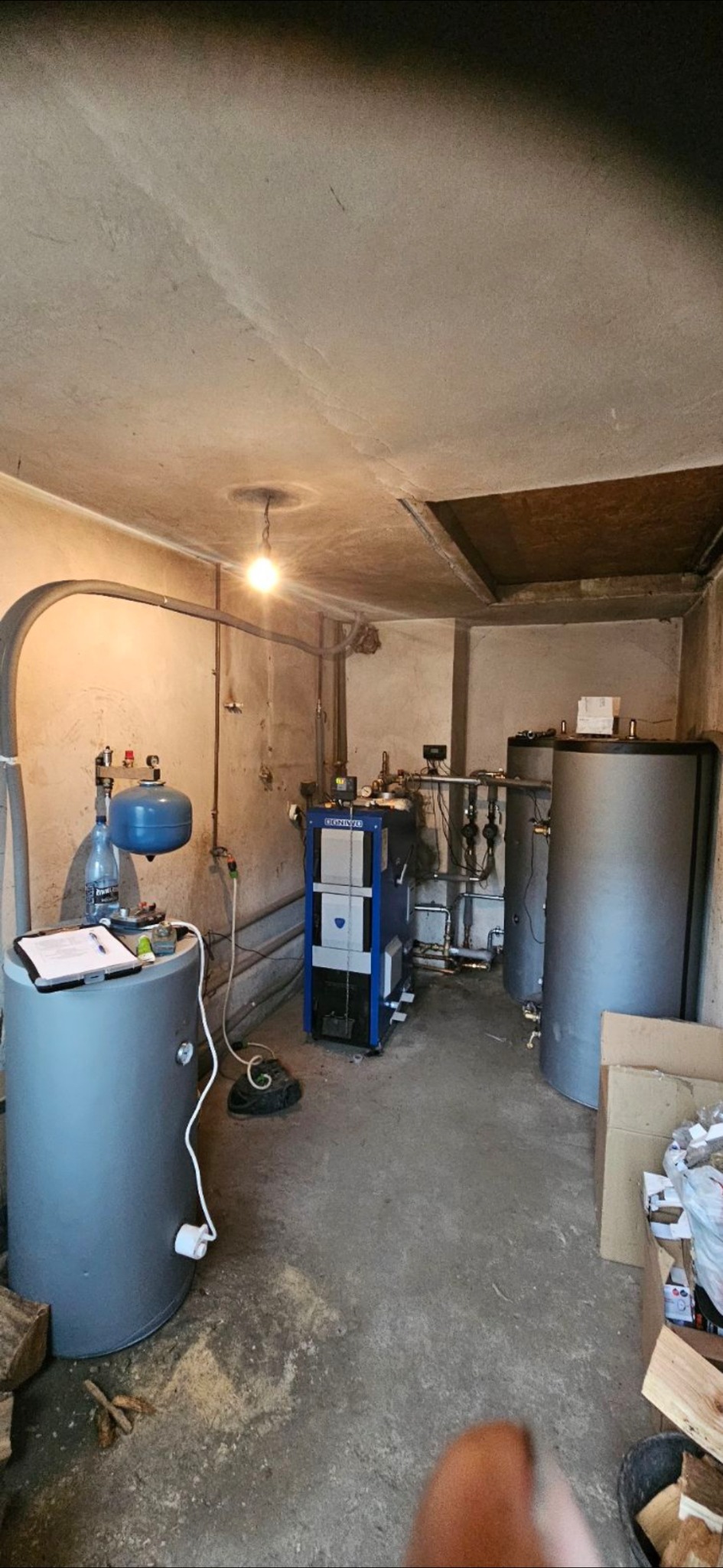 Kotłownia z piecem na paliwo stałe marki Ogniwo, zbiornikami buforowymi i osprzętem. Widoczne rury, zawory i instalacja. Na zbiorniku notatki i butelka wody.