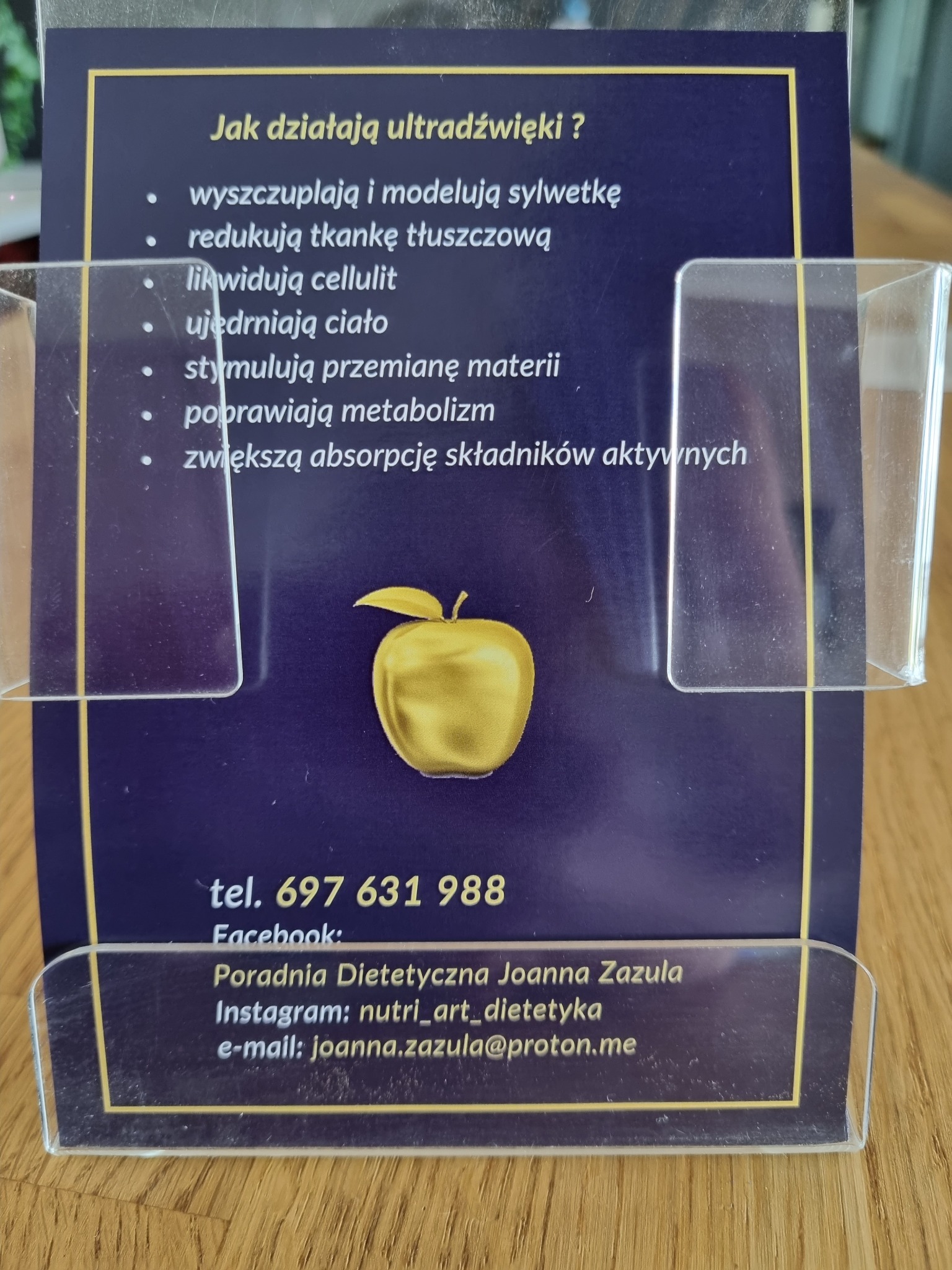 Ulotka informacyjna o działaniu ultradźwięków na ciało, z logo w kształcie złotego jabłka, umieszczona w przezroczystym stojaku. Zawiera informacje o wyszczuplaniu, redukcji tkanki tłuszczowej...