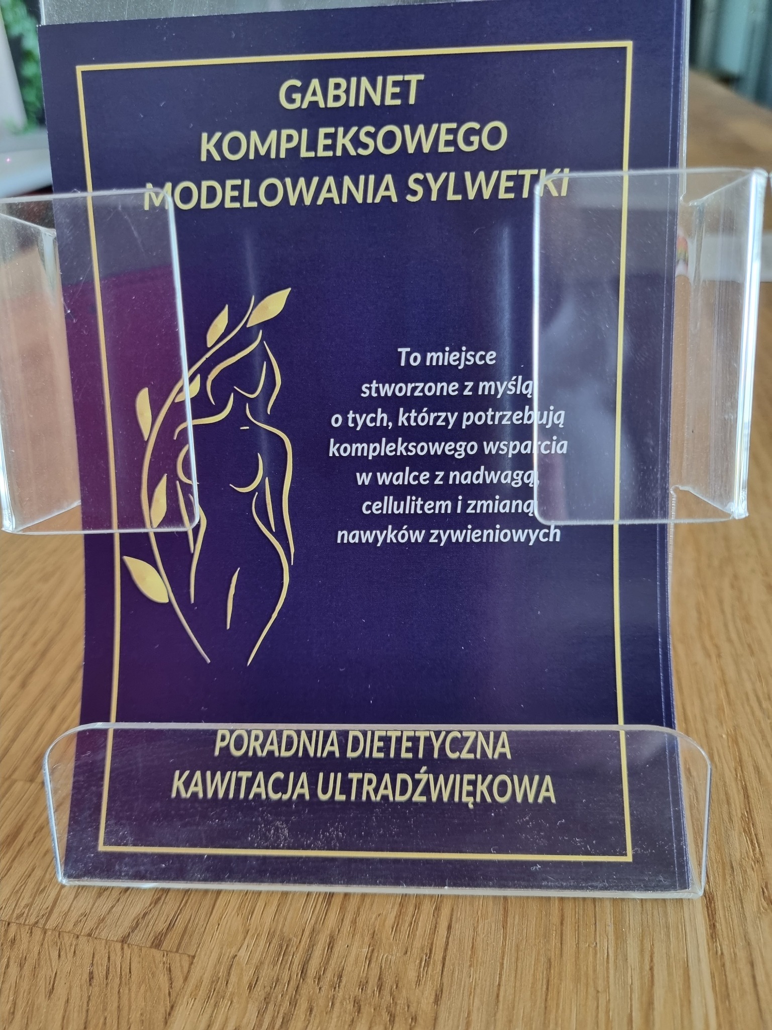 Ulotka reklamowa gabinetu kompleksowego modelowania sylwetki z poradnią dietetyczną i ofertą kawitacji ultradźwiękowej, umieszczona w przezroczystym stojaku na drewnianym blacie.